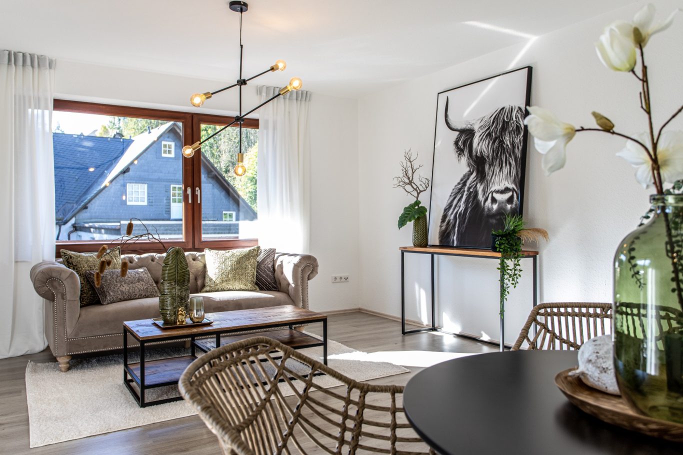 Immobilienmakler Münster Heller Wohnbereich mit Sofa, Tisch, Blumen, und einem großen Kunstwerk einer Kuh.